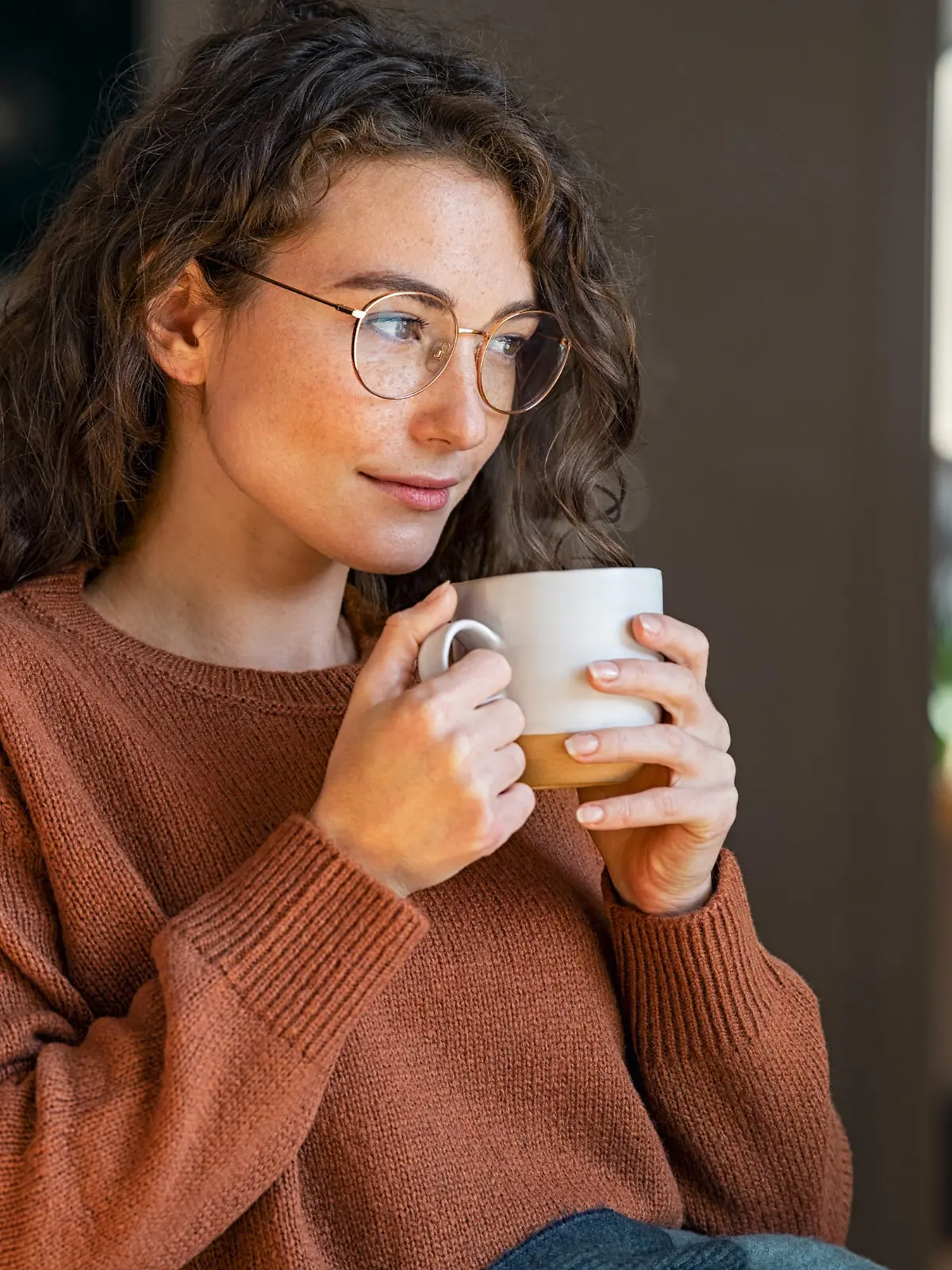 Pensive woman sitting in armchair and drinking hot coffee at home. Thoughtful young woman wearing eyeglasses drinking a cup of tea while thinking. Pretty contemplative girl with sweater relaxing at home while drinking purifying herbal tea during autumn, looking through the window.