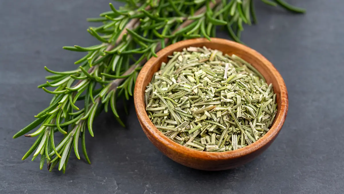 Fresh rosemary twigs and a wooden bowl with dried rosemary on a black slate background