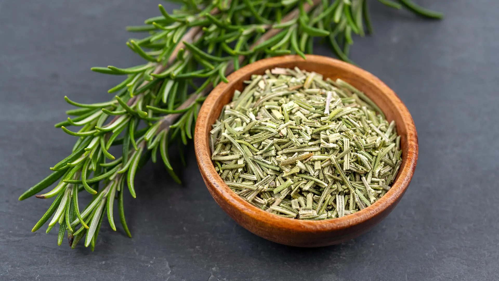 Fresh rosemary twigs and a wooden bowl with dried rosemary on a black slate background
