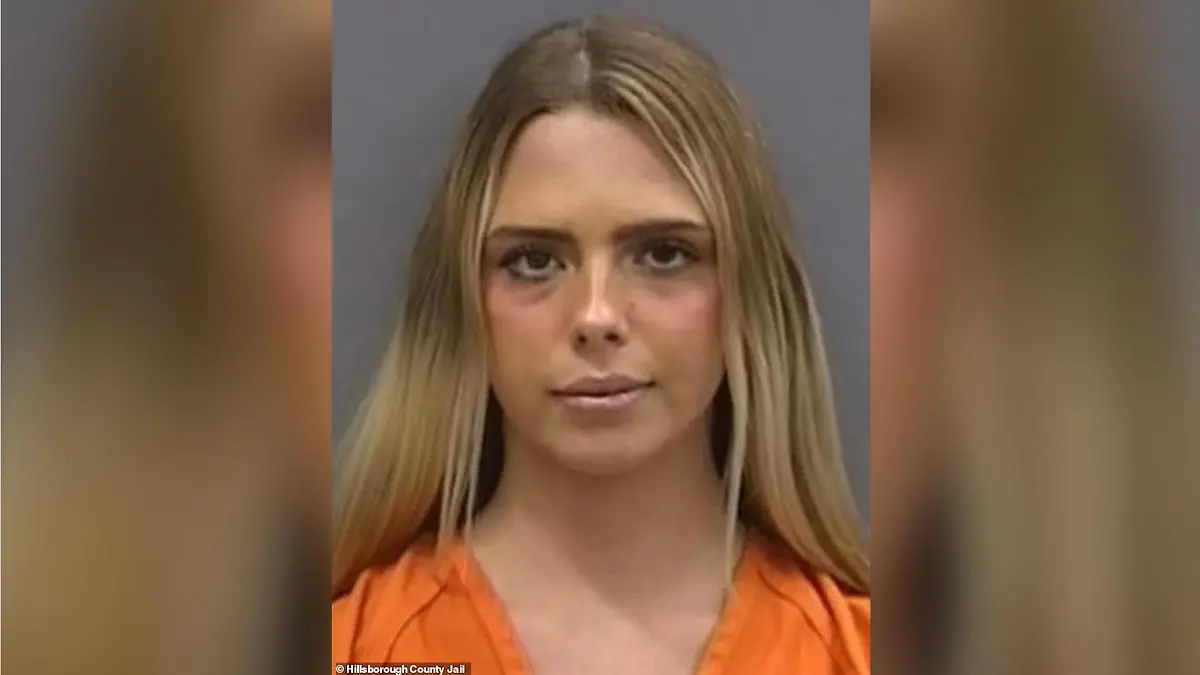 Eine blonde Frau in orangenem Oberteil steht vor einer grauen Wand und blickt direkt in die Kamera. Es handelt sich um ein Polizeifoto.