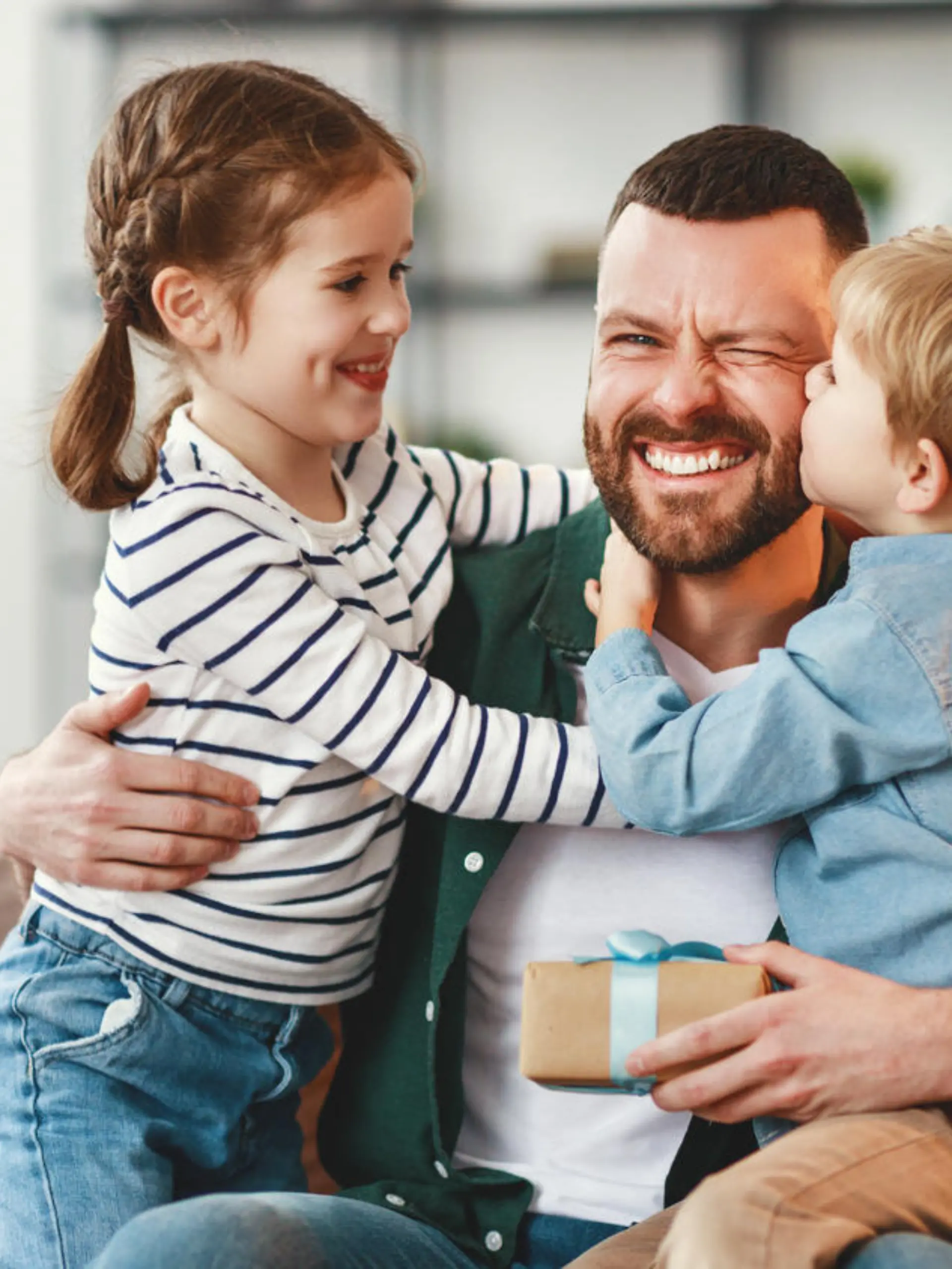 Joyful man with gift box in hand sitting on sofa at home and hugging cheerful son and daughter giving congratulations on fathers day