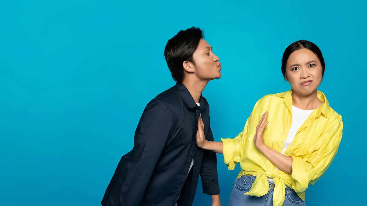 Asian man millennial puckering for a kiss towards a displeased woman who is rejecting the advance with a hand gesture, both against an expansive turquoise background, studio