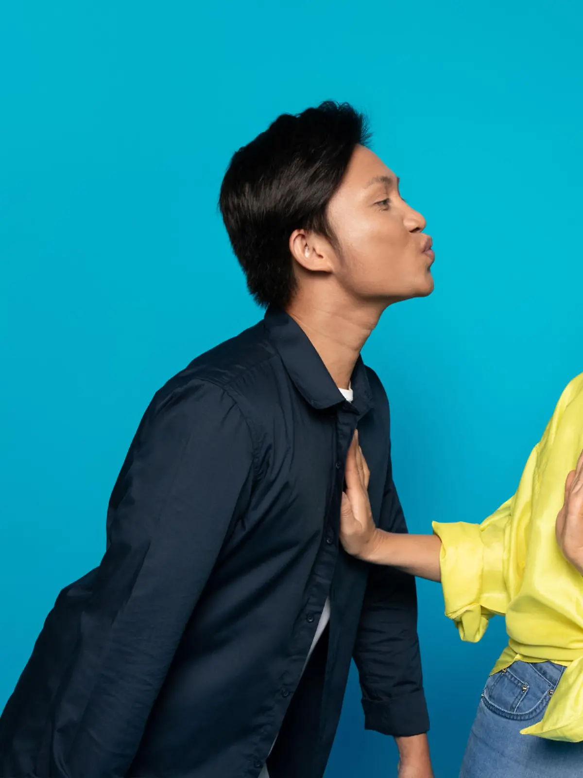 Asian man millennial puckering for a kiss towards a displeased woman who is rejecting the advance with a hand gesture, both against an expansive turquoise background, studio