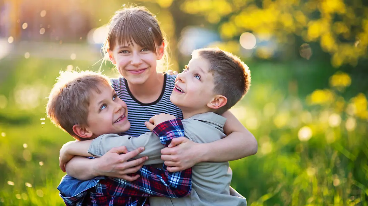 Little girl is embracing her younger brothers. The kids are playing in spring dandelion field. The girls is aged 10 and the boys are aged 6. They are enjoying beautiful nature laughing happily.