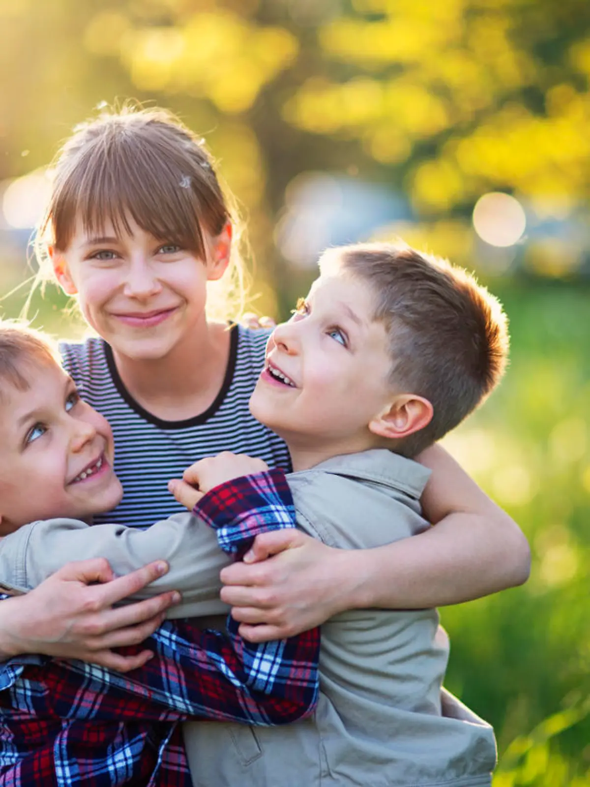 Little girl is embracing her younger brothers. The kids are playing in spring dandelion field. The girls is aged 10 and the boys are aged 6. They are enjoying beautiful nature laughing happily.
