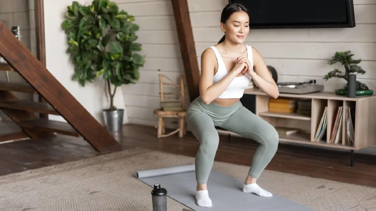 Young asian fitness girl doing squats workout at home, follow online sport gym instructor, standing on floor mat and smiling, doing exercises in living room.