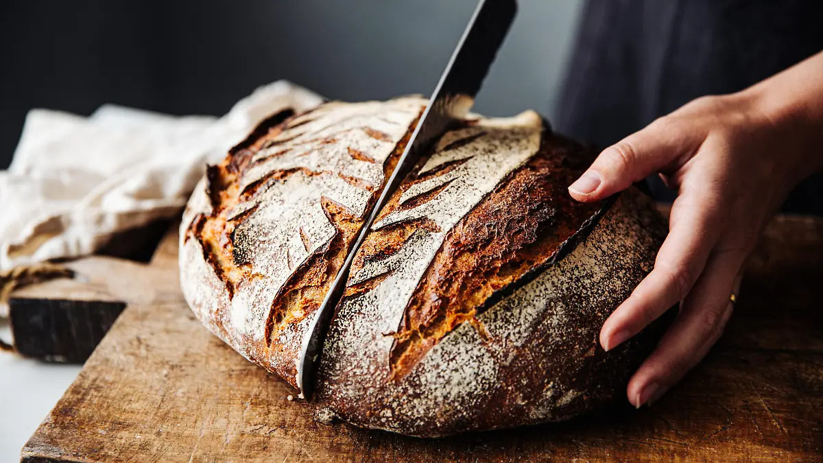 Close-up of woman cutting sourdough bread on board. Midsection of female is with knife and baked bread. She is in kitchen.