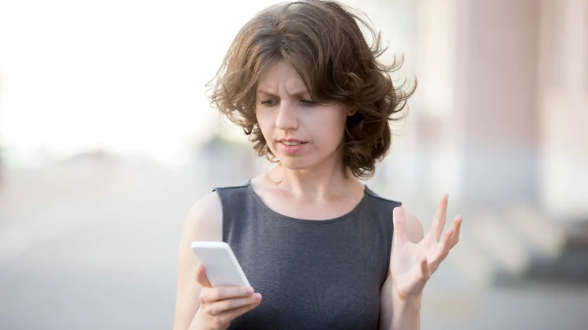Portrait of young woman holding cellphone in hands on the street in summer, looking at screen with irritated expression, made a mistake or annoyed by texts and calls