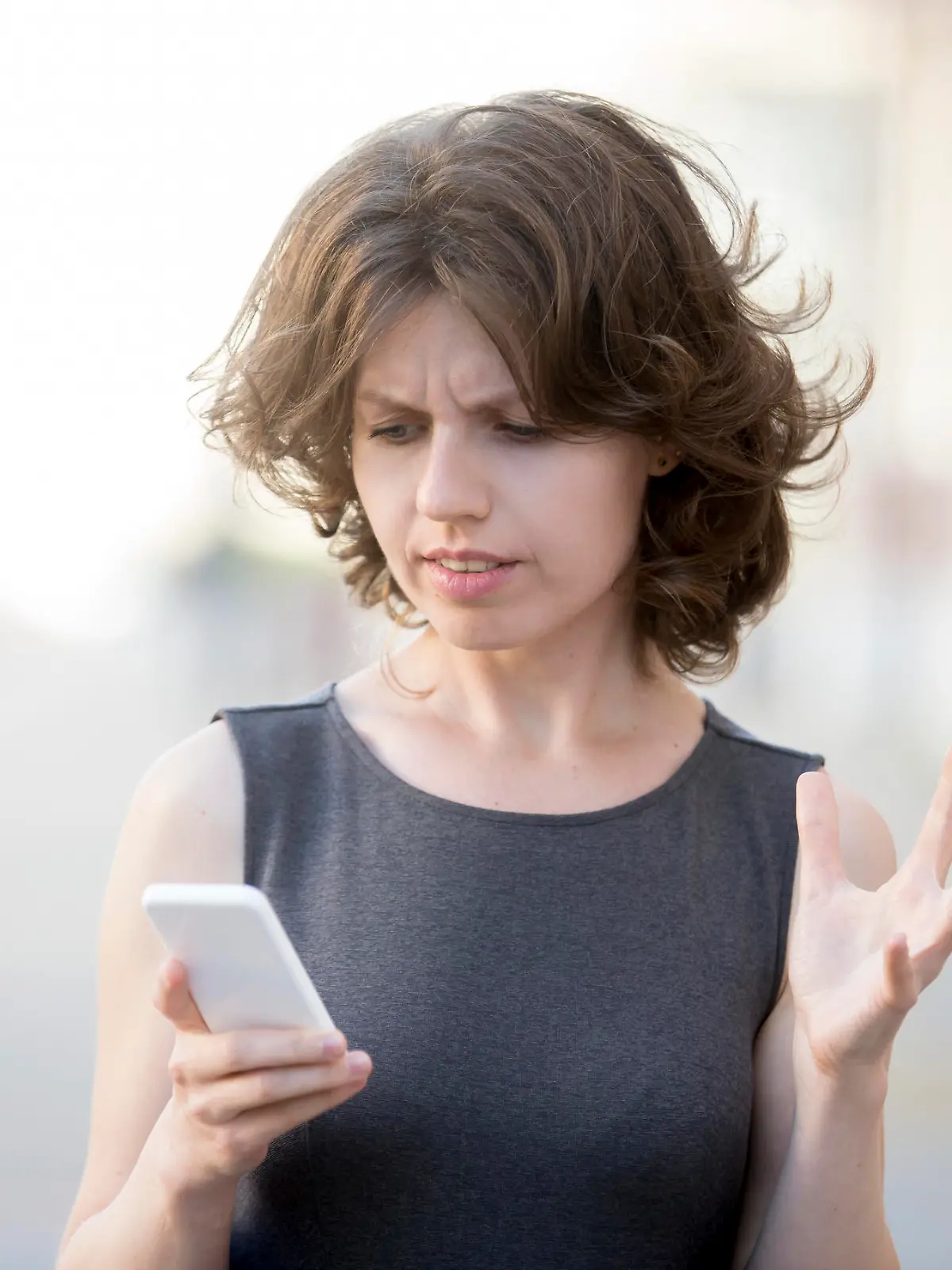 Portrait of young woman holding cellphone in hands on the street in summer, looking at screen with irritated expression, made a mistake or annoyed by texts and calls