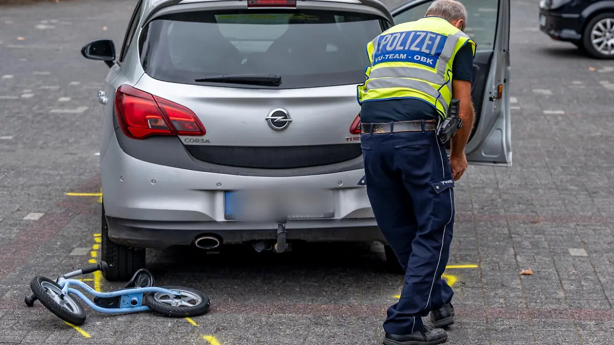 27.08.2022, Nordrhein-Westfalen, Marienheide: Mit Mitarbeiter der Polizei markiert die Unfallstelle auf einem Parkplatz, wo ein Zweijähriger von einem Auto angefahren und schwer verletzt worden war. Der 88 Jahre alte Autofahrer habe den Jungen am Samstagmittag bei der Suche nach einem Parkplatz übersehen, erklärte die Polizei. Das Kind sei auf dem Gelände vor einer Bankfiliale mit einem Laufrad unterwegs gewesen. Der Zweijährige kam nach notärztlicher Erstversorgung mit einem Rettungshubschrauber in eine Spezialklinik. Foto: Markus Klümper/dpa - ACHTUNG: Das Kfz-Zeichen wurde aus rechtlichen Gründen gepixelt +++ dpa-Bildfunk +++