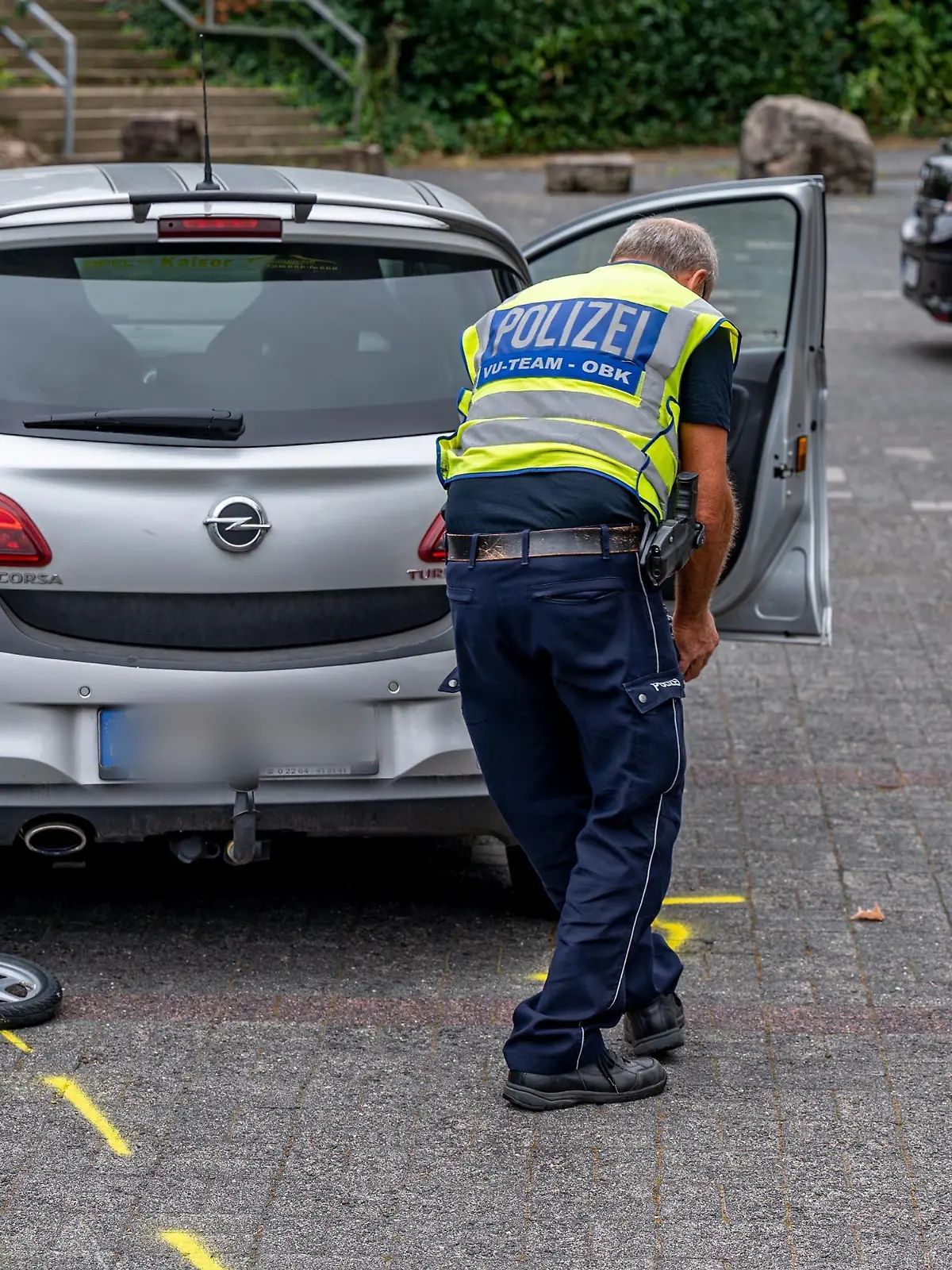 27.08.2022, Nordrhein-Westfalen, Marienheide: Mit Mitarbeiter der Polizei markiert die Unfallstelle auf einem Parkplatz, wo ein Zweijähriger von einem Auto angefahren und schwer verletzt worden war. Der 88 Jahre alte Autofahrer habe den Jungen am Samstagmittag bei der Suche nach einem Parkplatz übersehen, erklärte die Polizei. Das Kind sei auf dem Gelände vor einer Bankfiliale mit einem Laufrad unterwegs gewesen. Der Zweijährige kam nach notärztlicher Erstversorgung mit einem Rettungshubschrauber in eine Spezialklinik. Foto: Markus Klümper/dpa - ACHTUNG: Das Kfz-Zeichen wurde aus rechtlichen Gründen gepixelt +++ dpa-Bildfunk +++