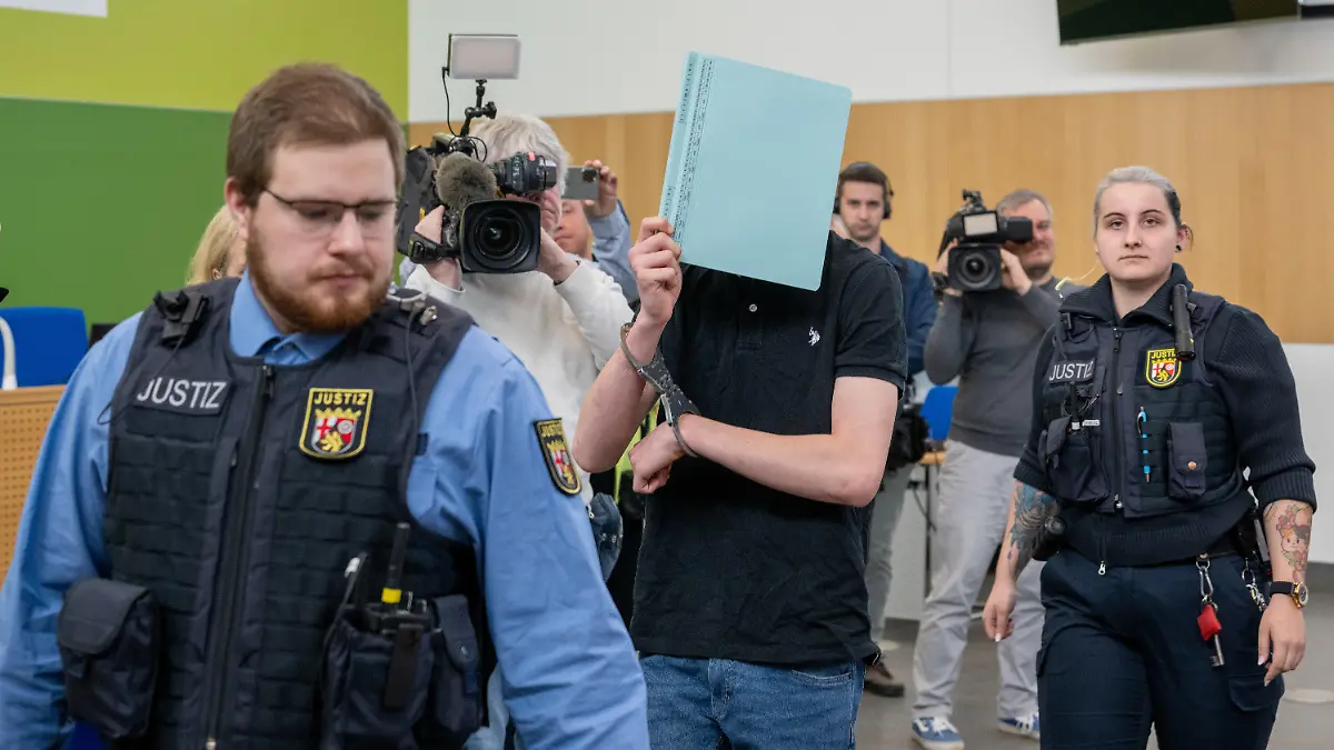 15.04.2024, Rheinland-Pfalz, Trier: Ein Angeklagter (M) wird wegen gemeinschaftlichen Mord an einem Gerolsteiner Arzt von Justizbediensteten zur Anklagebank geführt. Er soll mit einem Bekannten und seiner Mutter einen Arzt aus der Eifel getötet haben. Foto: Harald Tittel/dpa +++ dpa-Bildfunk +++