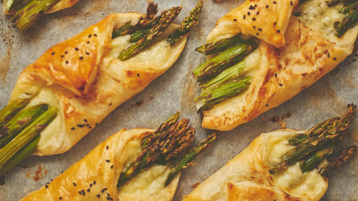 Baked green asparagus in puff pastry sprinkled with sesame seeds and cheese. Placed on a white baking paper background. Close-up, top view, flat lay.