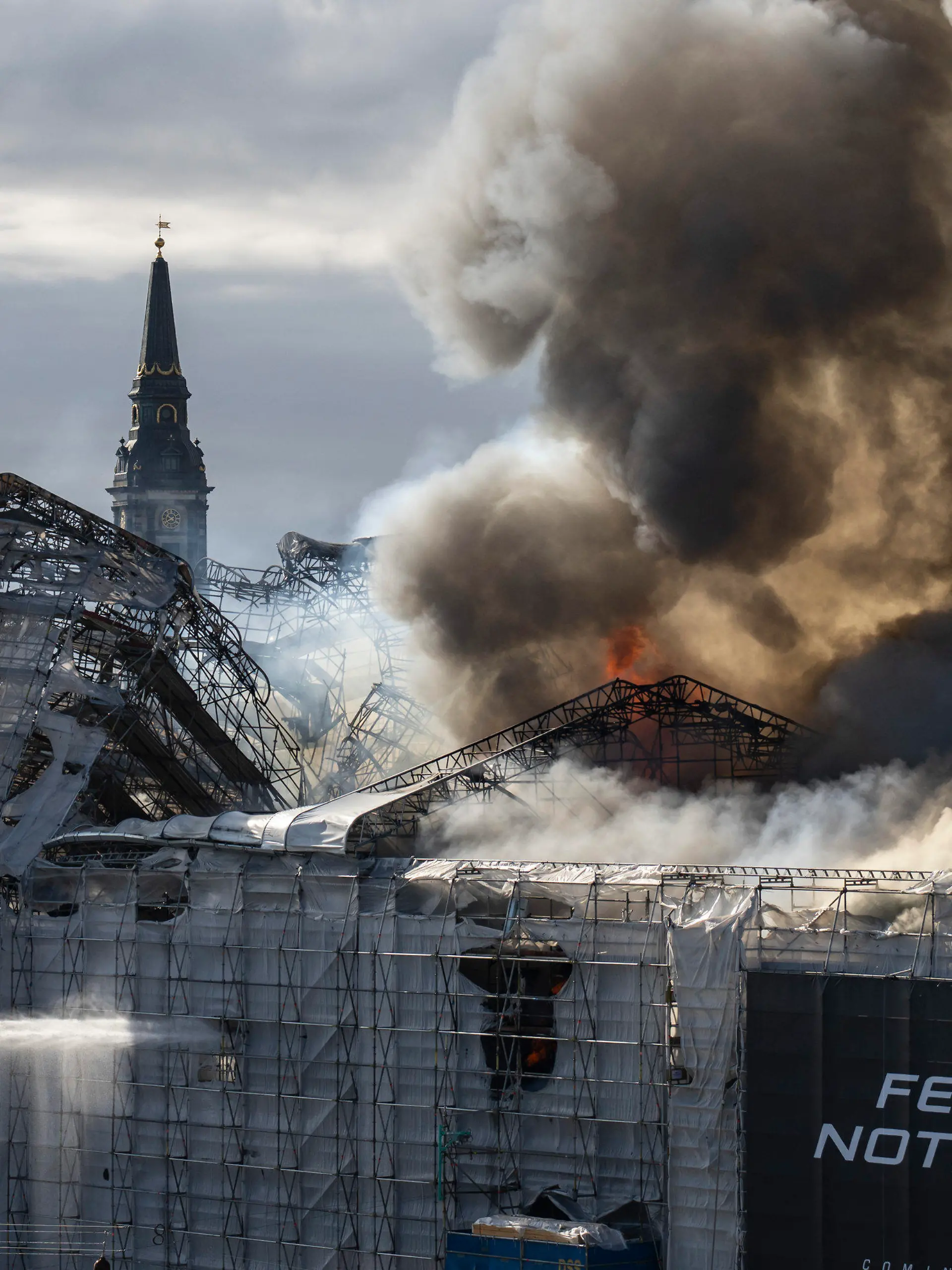 16.04.2024, Dänemark, Kopenhagen: Feuerwehrleute arbeiten neben dem Schriftzug «Fear nothing» (fürchte nichts), während Rauch aus der Alten Börse («Boersen») aufsteigt. Eines der ältesten Gebäude Kopenhagens steht in Flammen, und seine ikonische Turmspitze ist eingestürzt. Das Dach der alten Börse (Boersen) aus dem 17. Jahrhundert, die einst das Finanzzentrum Dänemarks war, wurde am Dienstag von Flammen zerstört. Foto: Emil Nicolai Helms/Ritzau Scanpix Foto/AP +++ dpa-Bildfunk +++