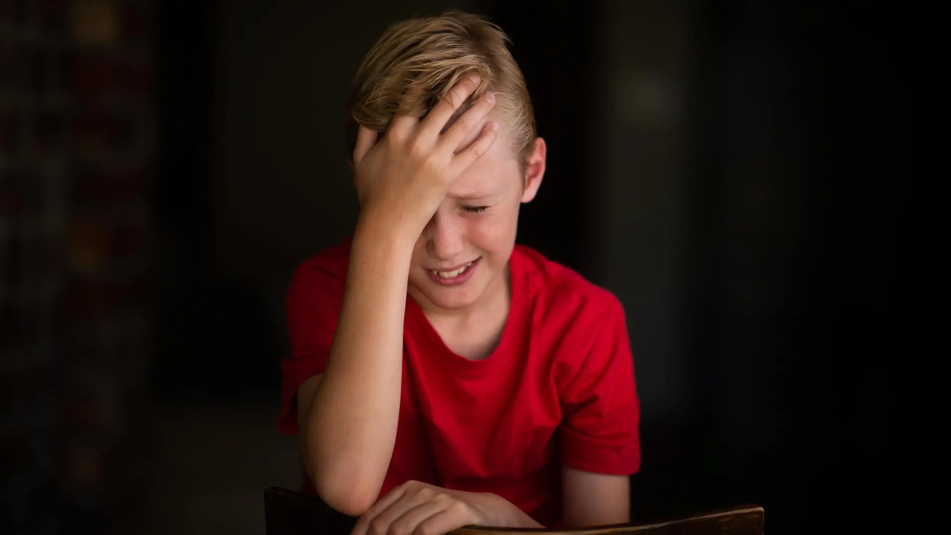 A young 11-year old boy sitting on a chair with his hand touching his head. His face is showing pain or frustration. He is looking sad.
