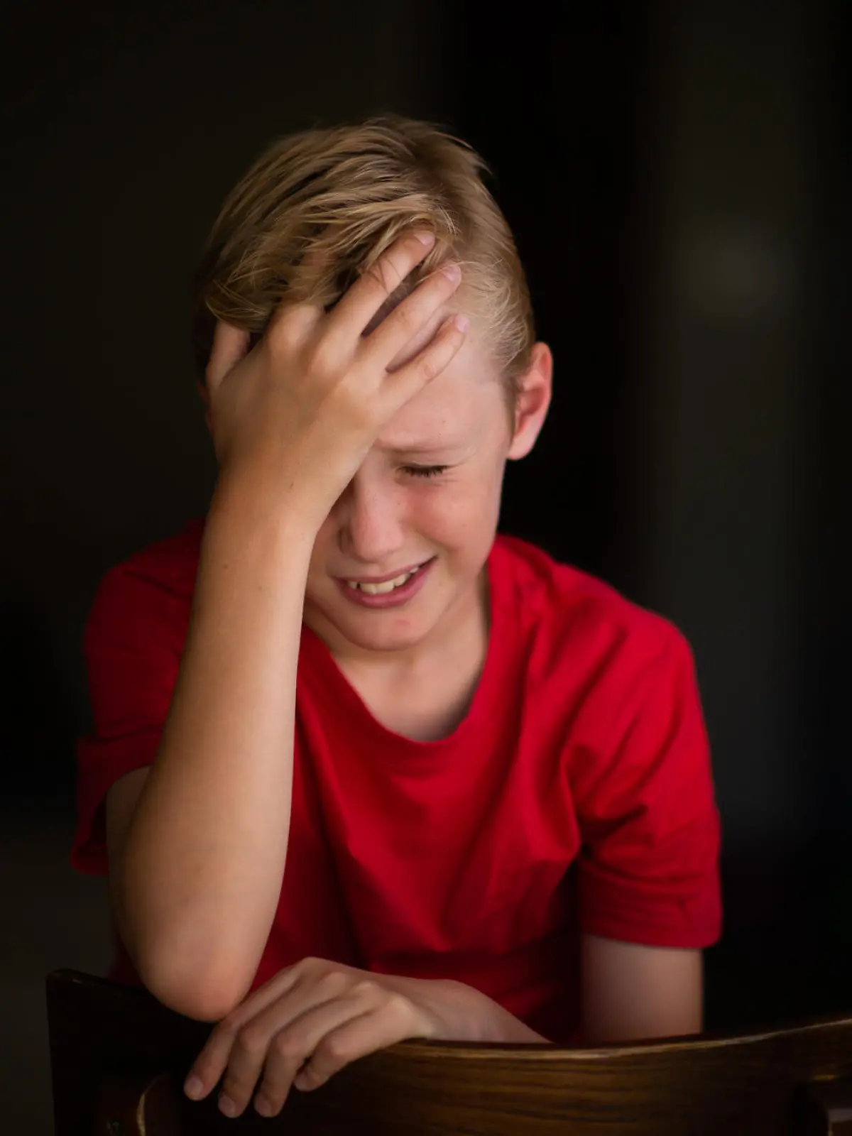 A young 11-year old boy sitting on a chair with his hand touching his head.  His face is showing pain or frustration.  He is looking sad.