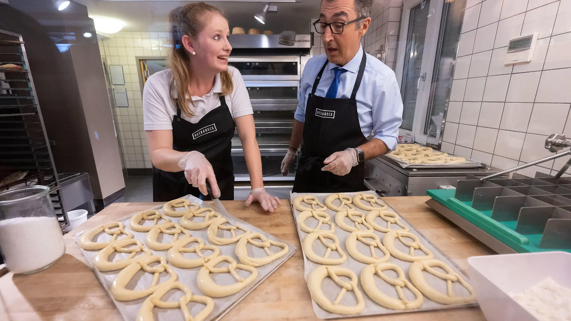 25.08.2022, Baden-Württemberg, Bad Urach: Bundesernährungsminister Cem Özdemir (Bündnis 90/Die Grünen) und Luisa Beck, die Tochter des Inhabers der Großbäckerei BeckaBeck, formen während des Besuchs einer Filiale der Bäckerei Brezeln. Foto: Marijan Murat/dpa +++ dpa-Bildfunk +++