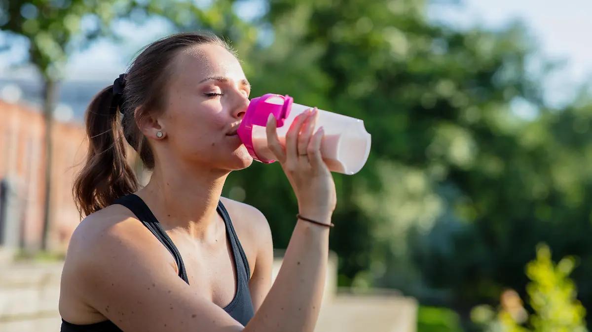 A young woman is sitting outdoors in work out clothes. She is holding a protein shake.