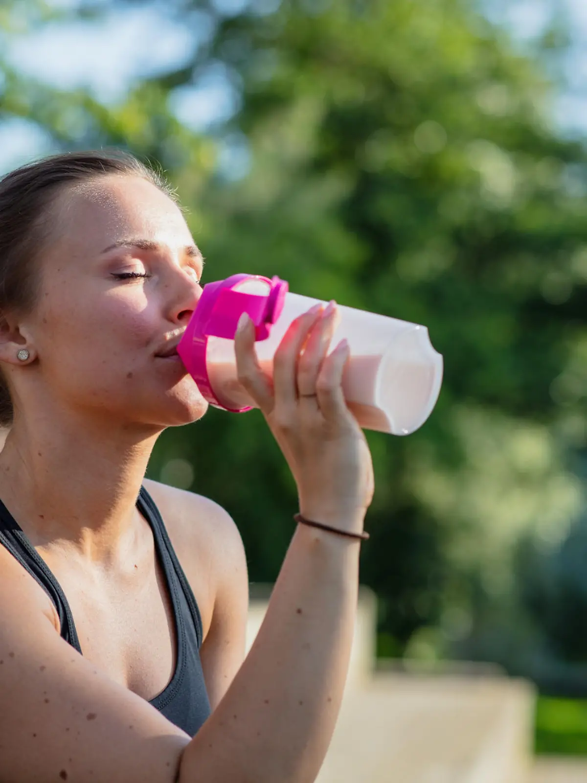 A young woman is sitting outdoors in work out clothes. She is holding a protein shake.