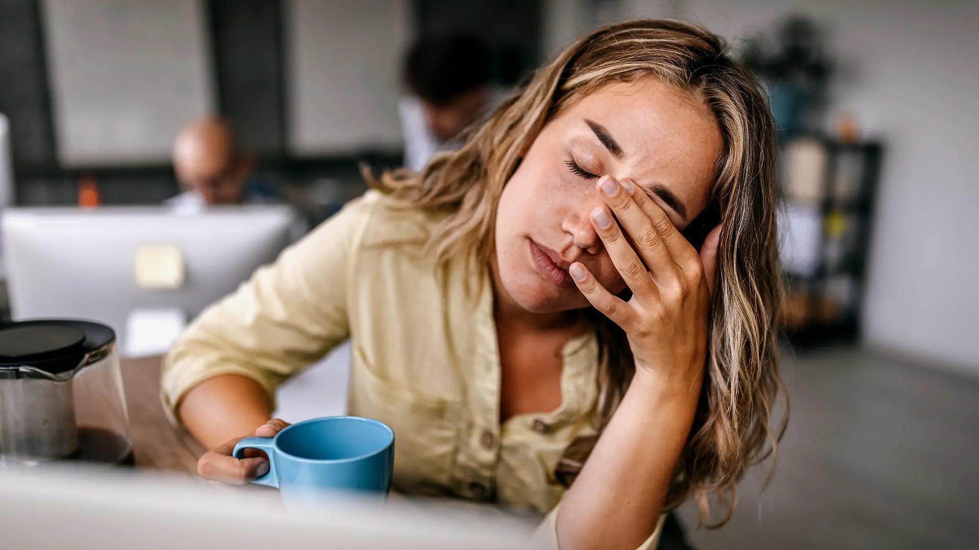 Tired and exhausted young female entrepreneur drinking coffee and rubbing eyes while feeling stressed and worried in coworking space