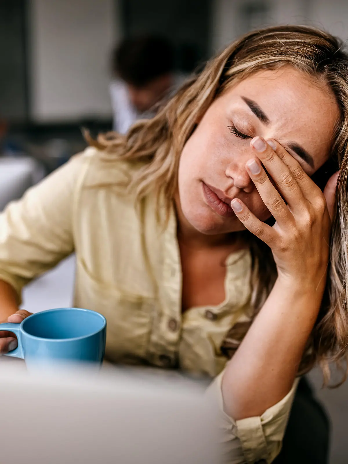 Tired and exhausted young female entrepreneur drinking coffee and rubbing eyes while feeling stressed and worried in coworking space