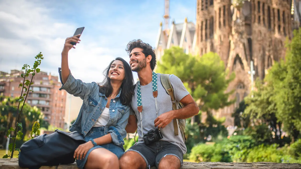 Male and female travelers sitting on wall in public park near Sagrada Familia in Barcelona and taking selfie on sunny summer day.