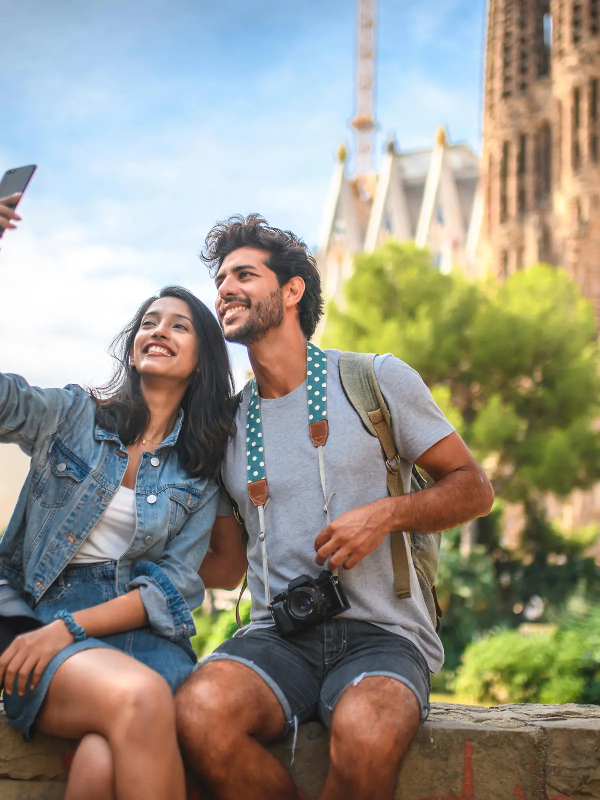 Male and female travelers sitting on wall in public park near Sagrada Familia in Barcelona and taking selfie on sunny summer day.