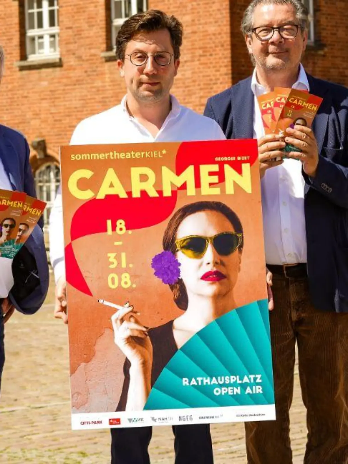 Die Premiere der Kieler Sommeroper „Carmen“ auf dem Rathausplatz. Die Theaterleitung mit dem Carmen-Plakat.