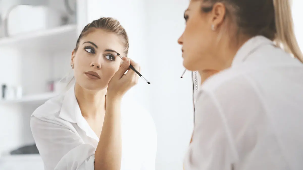 Closeup rear view of attractive mid 20's blond woman putting on some makeup before heading to work. She's in front of large bathroom mirror, focus is on her reflection.