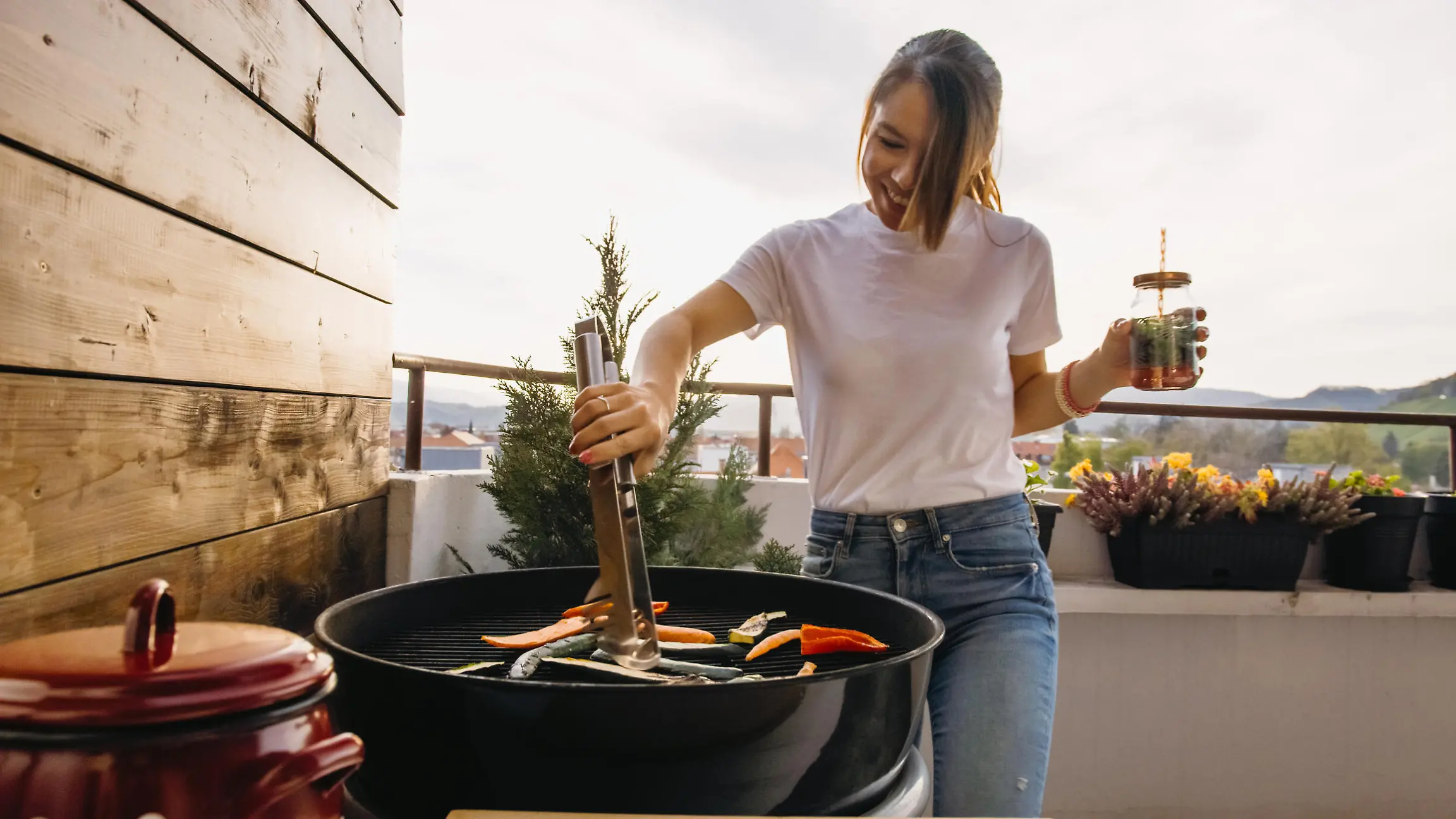 Smiling young woman using tongs to grill food on a barbecue while drinking on the terrace