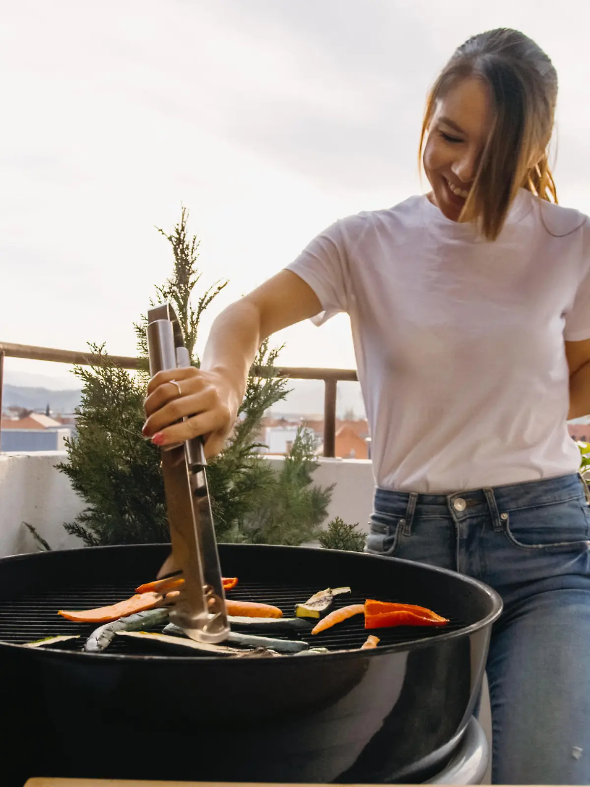 Smiling young woman using tongs to grill food on a barbecue while drinking on the terrace