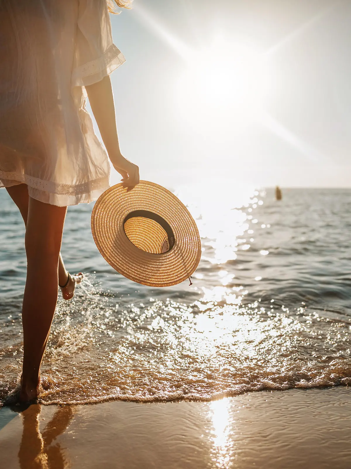 Close-up of young woman in white sun dress and with hat in hand walking alone on sandy beach at summer sunset, splashing water in sea shallow