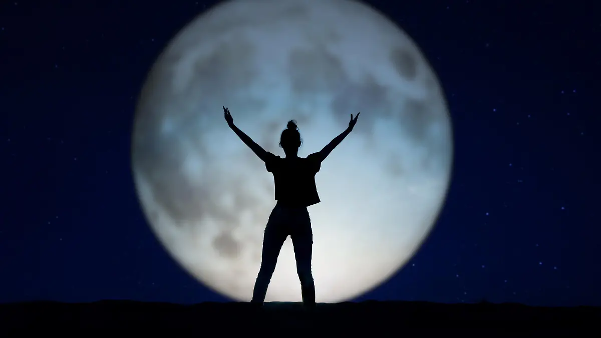 Silhouette of a woman standing in the night with the arms up, giant moon in the background