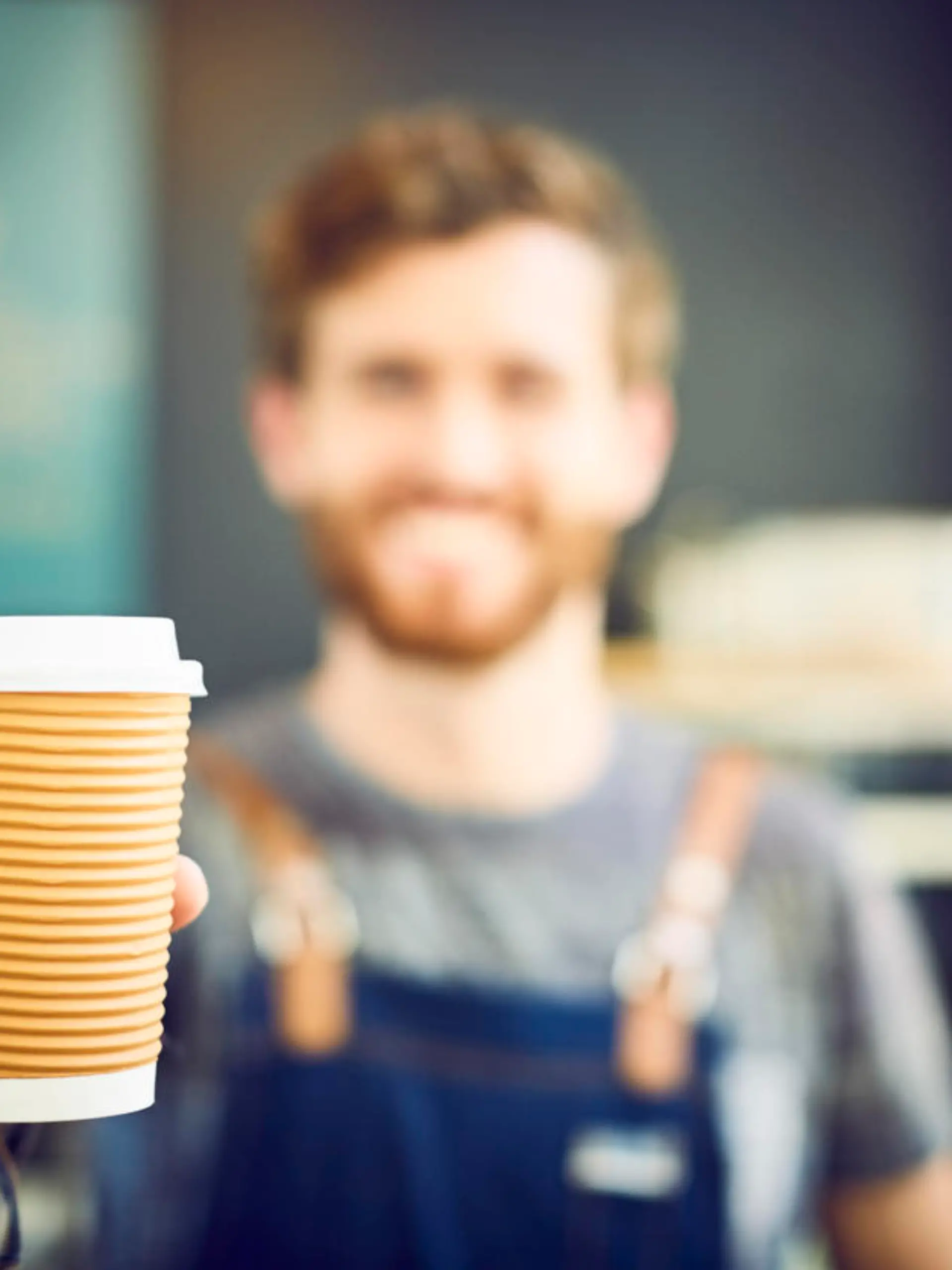 Young barista offering coffee in disposable cup. Smiling waiter is serving drink in coffee shop. Focus is on container.