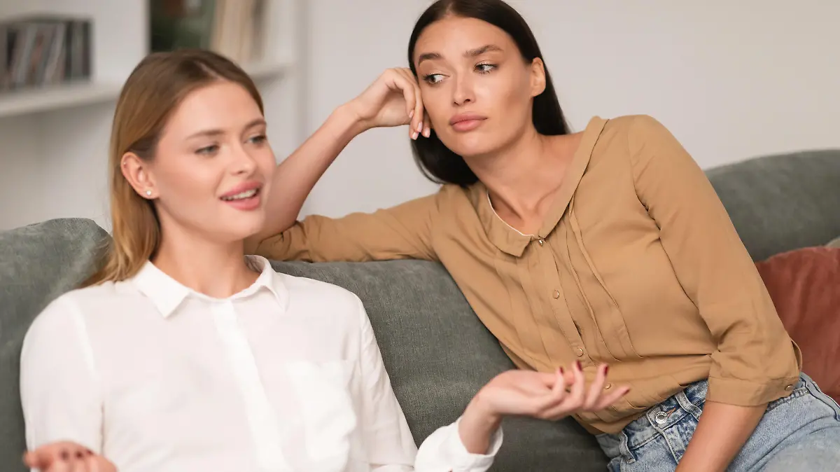 Jealous Woman Listening With Envy To Her Friend Bragging About Great Life Sitting On Sofa In Modern Living Room At Home. Envious Lousy Friends, Fake Friendship Concept. Selective Focus