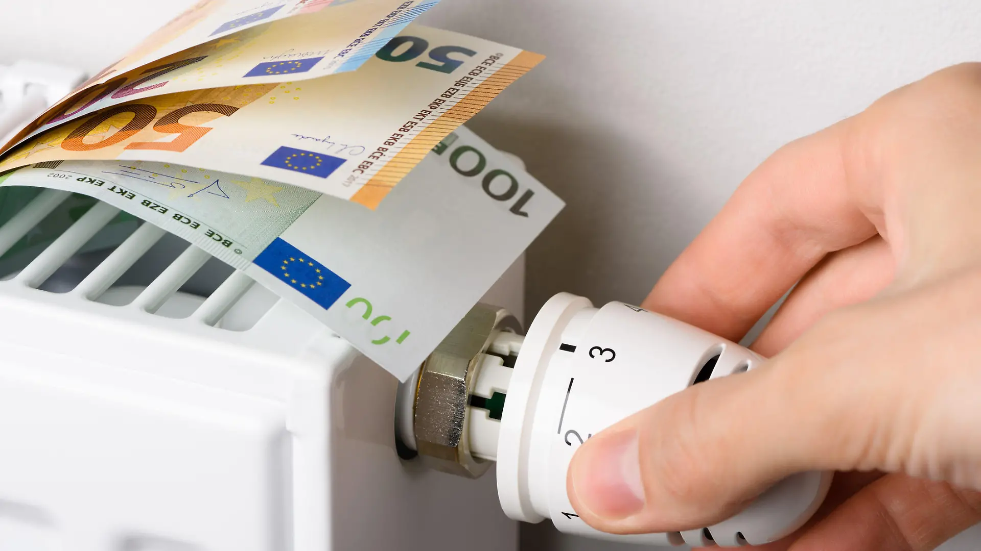 Hand adjusting the valve knob of heating radiator temperature thermostat with of euro money banknotes in it. Expensive heating costs and rising energy bill prices for winter cold season