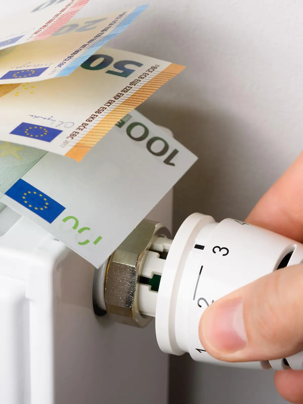 Hand adjusting the valve knob of heating radiator temperature thermostat with of euro money banknotes in it. Expensive heating costs and rising energy bill prices for winter cold season