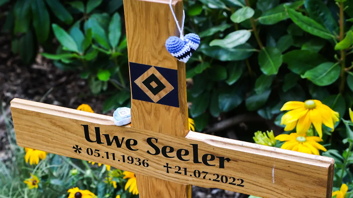 09.08.2022, Hamburg: Ein Holzkreuz steht auf dem Grab der Fußballlegende Uwe Seeler, an dem Kreuz hängt ein kleines gehäkeltes Herz in den Farben des HSV. Der beliebte Sportler wurde am 04. August im engsten Familienkreis auf dem Friedhof Ohlsdorf beigesetzt. Um den vielen Menschen die Suche nach dem Grab zu erleichtern, hat die Friedhofsverwaltung die kleinen Wegbeschreibungen zur Verfügung gestellt. (zu dpa "Große Feier für großartigen Menschen: Abschied von Uwe Seeler") Foto: Christian Charisius/dpa +++ dpa-Bildfunk +++