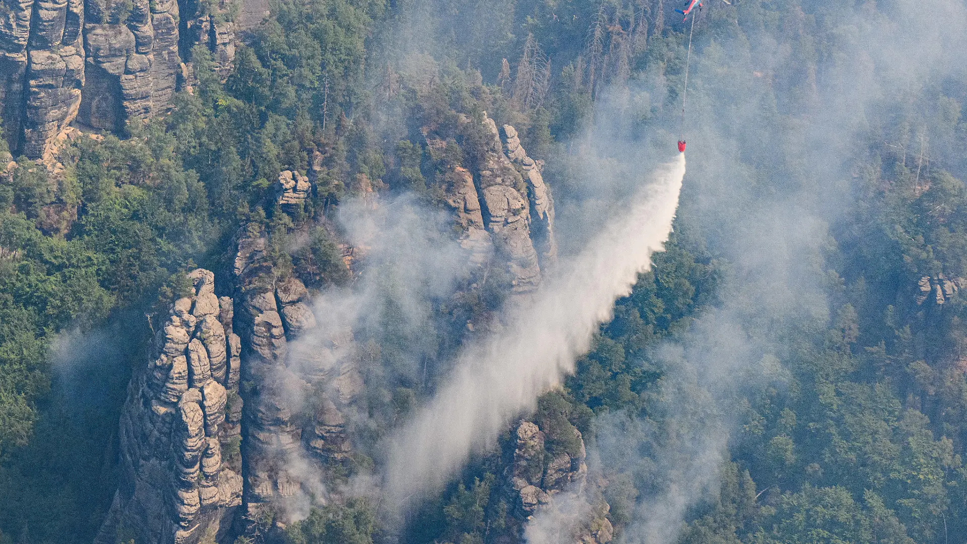 28.07.2022, Sachsen, Schmilka: Ein Lastenhubschrauber aus Österreich fliegt mit einem Löschwasser-Außenlastbehälter um einen Waldbrand im Nationalpark Sächsische Schweiz zu löschen. Foto: Robert Michael/dpa +++ dpa-Bildfunk +++