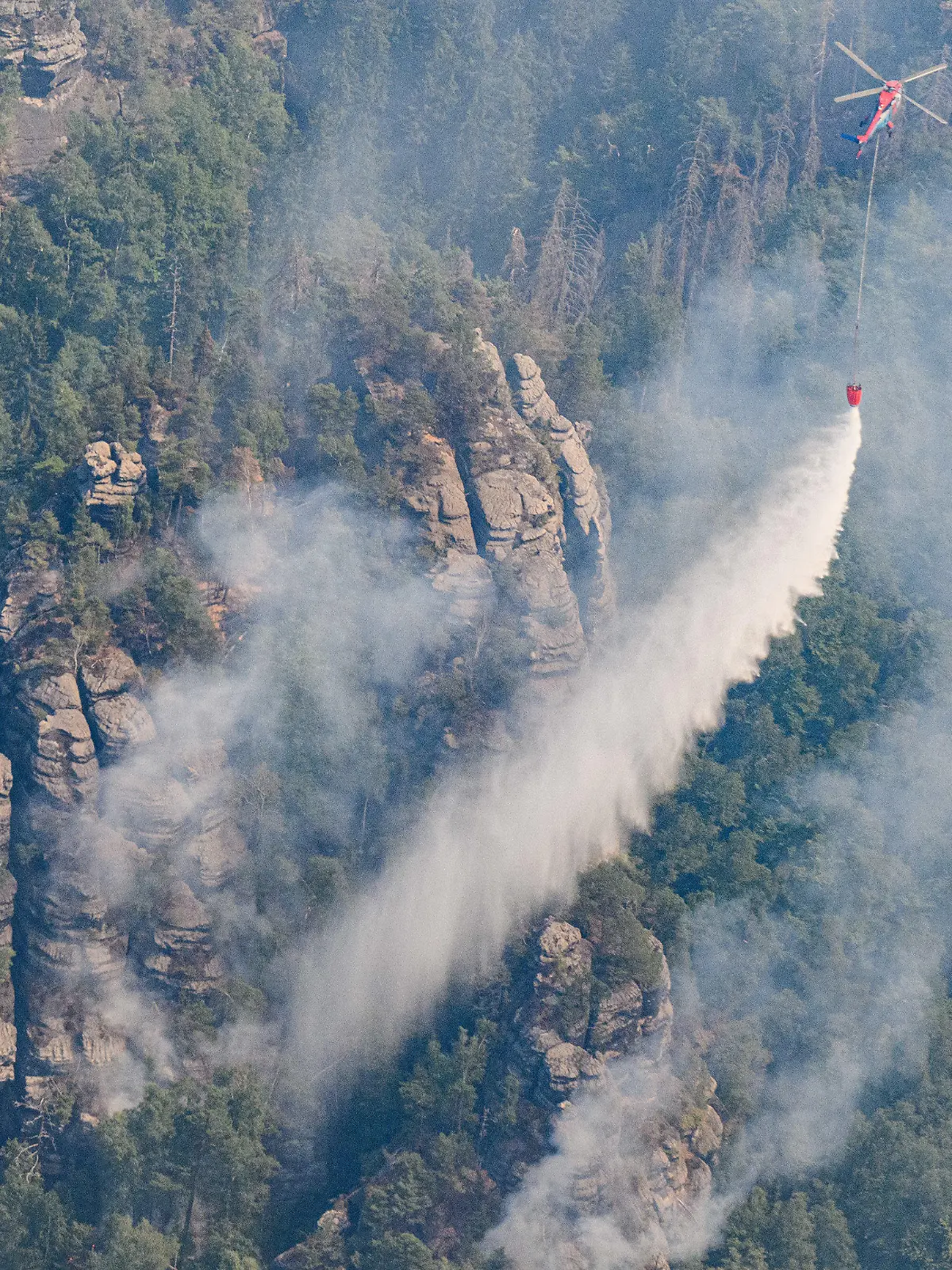 28.07.2022, Sachsen, Schmilka: Ein Lastenhubschrauber aus Österreich fliegt mit einem Löschwasser-Außenlastbehälter um einen Waldbrand im Nationalpark Sächsische Schweiz zu löschen. Foto: Robert Michael/dpa +++ dpa-Bildfunk +++