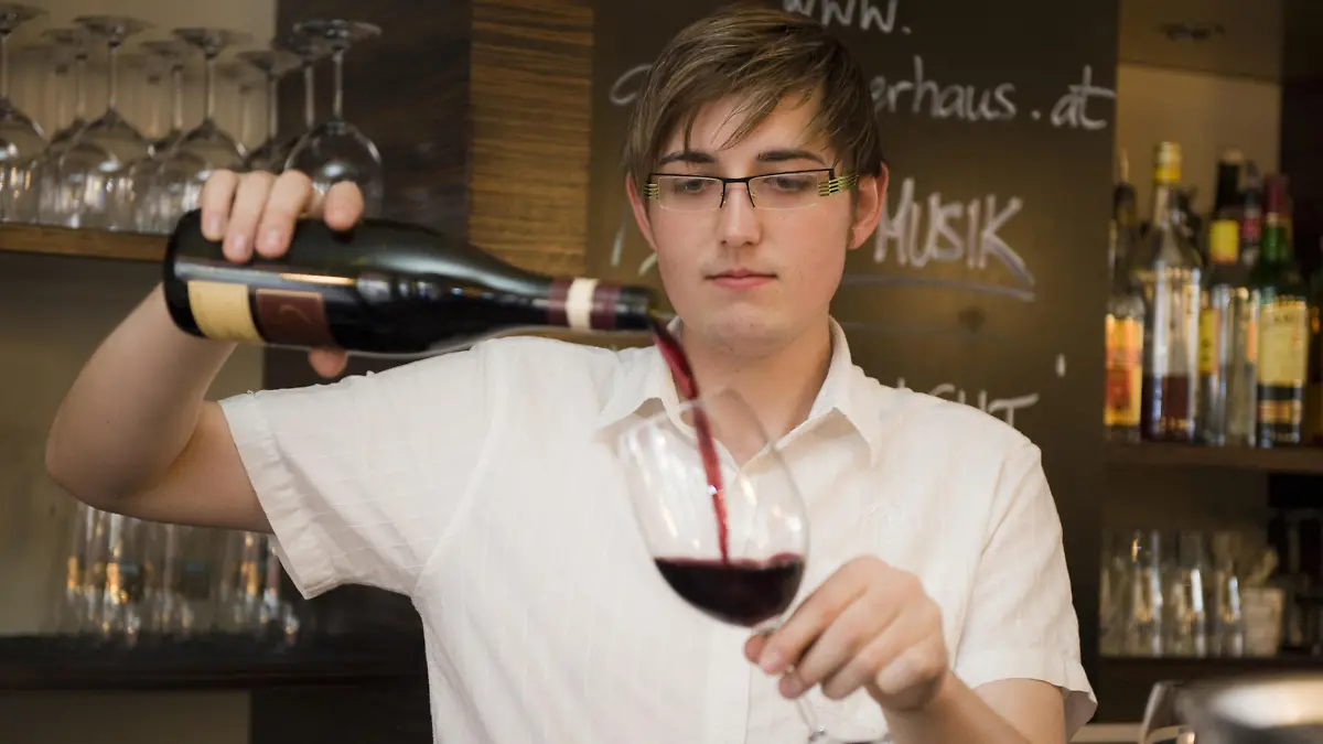 Portraet eines jungen, sympathischen Kellner in einem Restaurant, ein Weinglas fuellend | Portrait of a young likeable waiter in a Restaurant filling a glass of wine