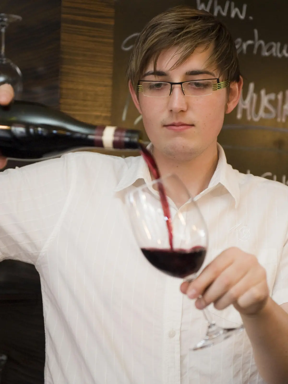 Portraet eines jungen, sympathischen Kellner in einem Restaurant, ein Weinglas fuellend | Portrait of a young likeable waiter in a Restaurant filling a glass of wine