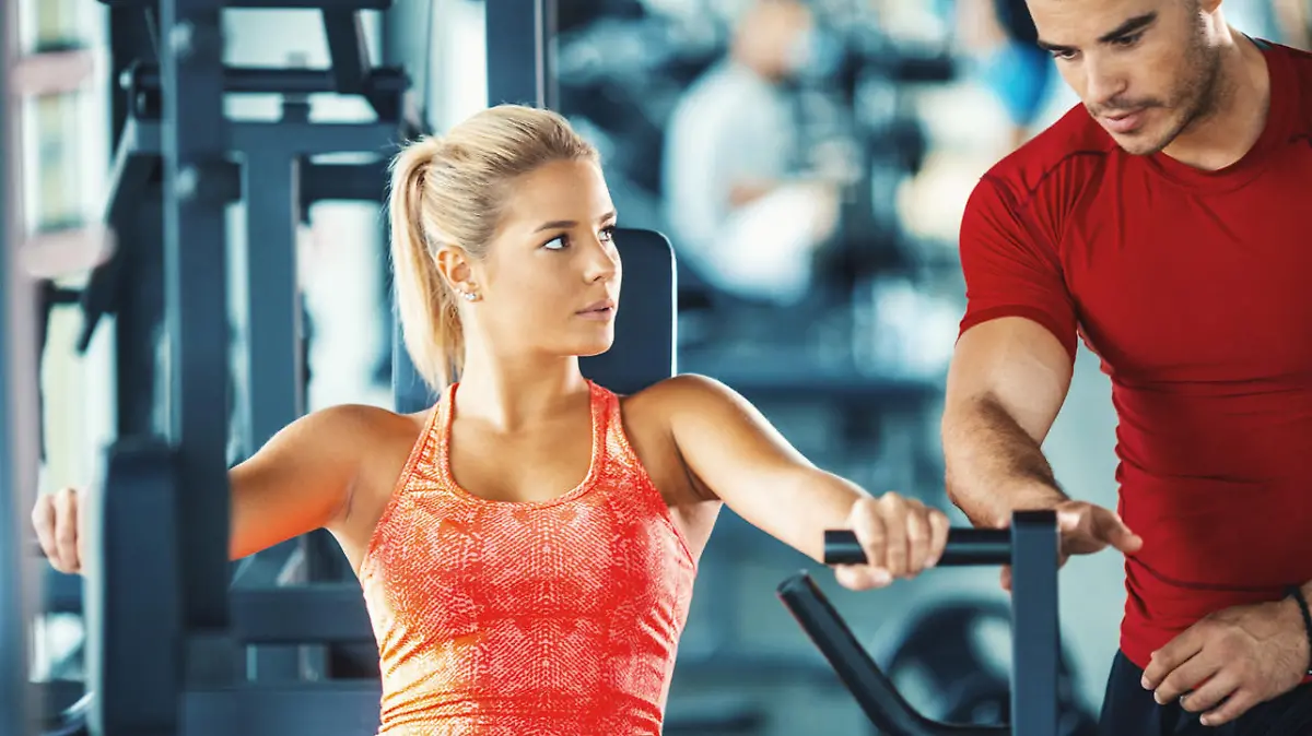 Closeup front view of mid 20's blond woman working out on a chest press machine. Her instructor is standing next to her and making sure she's doing it properly.