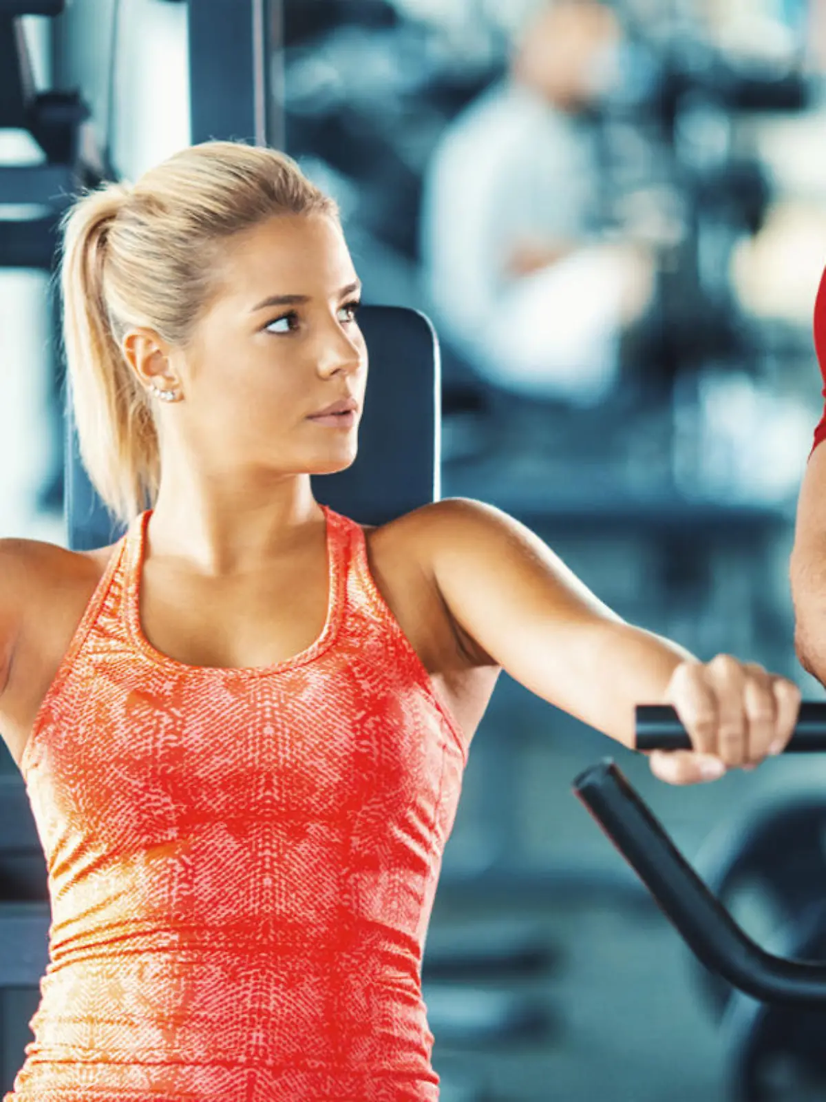 Closeup front view of mid 20's blond woman working out on a chest press machine. Her instructor is standing next to her and making sure she's doing it properly.