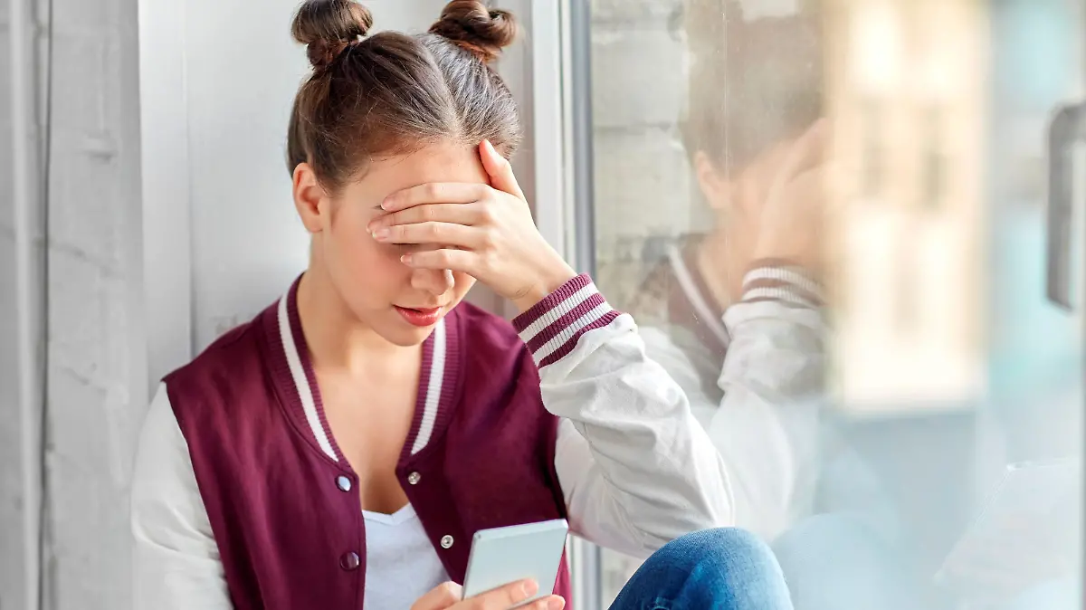 technology, cyberbullying and people concept - sad teenage girl with smartphone sitting on window sill