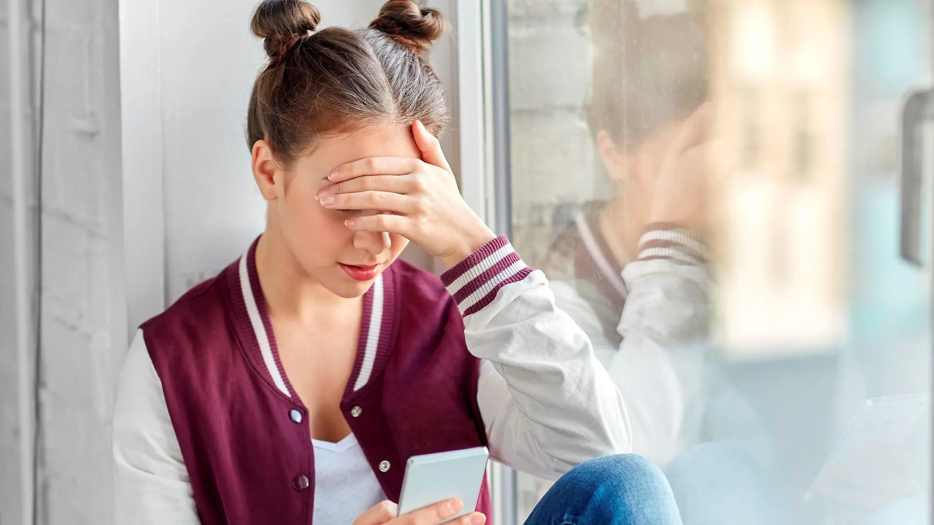 technology, cyberbullying and people concept - sad teenage girl with smartphone sitting on window sill