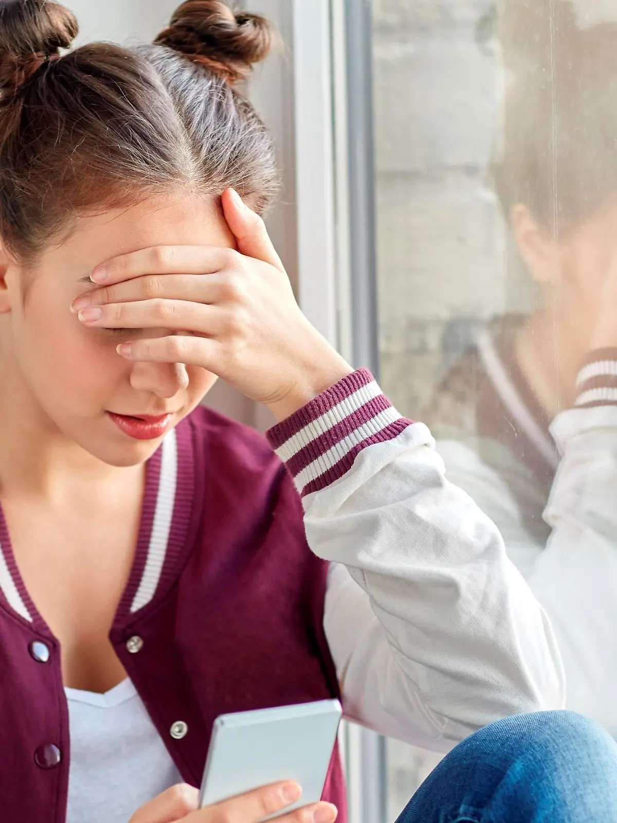 technology, cyberbullying and people concept - sad teenage girl with smartphone sitting on window sill