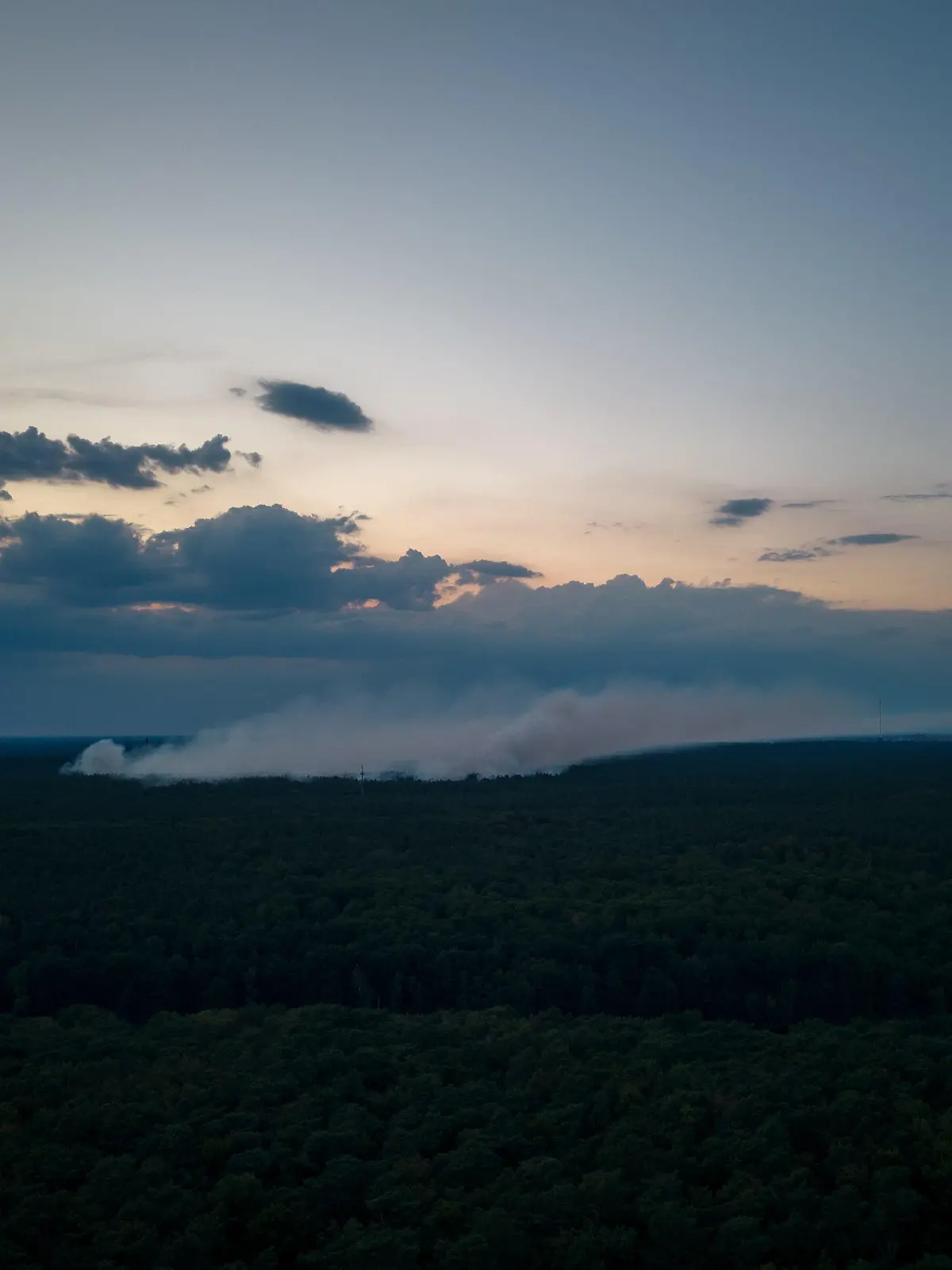 dpatopbilder - 04.08.2022, Berlin: Die Rauchwolke über der Brandstelle im Grunewald in der Dämmerung, aufgenommen aus der nahegelegen Waldsiedlung an der Krummen Lanke (Aufnahme mit Drohne). Am Morgen ist ein Feuer auf dem Sprengplatz der Polizei im Grunewald ausgebrochen. Durch den Brand ist es am frühen Abend erneut zu Explosionen gekommen. Foto: Christian Ender/dpa +++ dpa-Bildfunk +++
