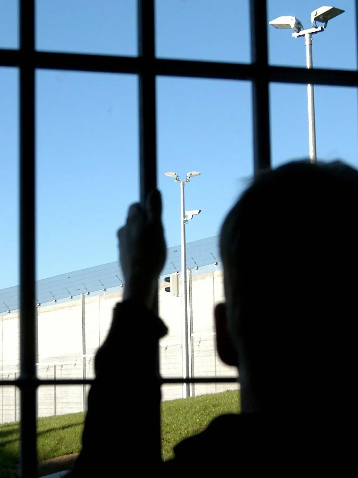 Ein Journalist steht mit einer Hand am Gitter einer Zelle der neuen Justizvollzugsanstalt im bayerischen Kempten am 4.9.2003. Er blickt auf Überwachungsanlagen mit Kameras und einer meterhohen Mauer mit Zaun. Wenige Wochen vor der Inbetriebnahme der JVA Kempten erhielten Journalisten die Möglichkeit, unter realitätsnahen Bedingungen eine Nacht hinter Gittern zu verbringen. Neben der obligatorischen Gefangenenkleidung mussten sie sich auch dem Tagesablauf in einer JVA unterordnen. Die JVA in Kempten mit 338 Haftplätzen wurde für mehr als 40 Millionen Euro errichtet und nimmt ab Oktober ihren regulären Betrieb auf.