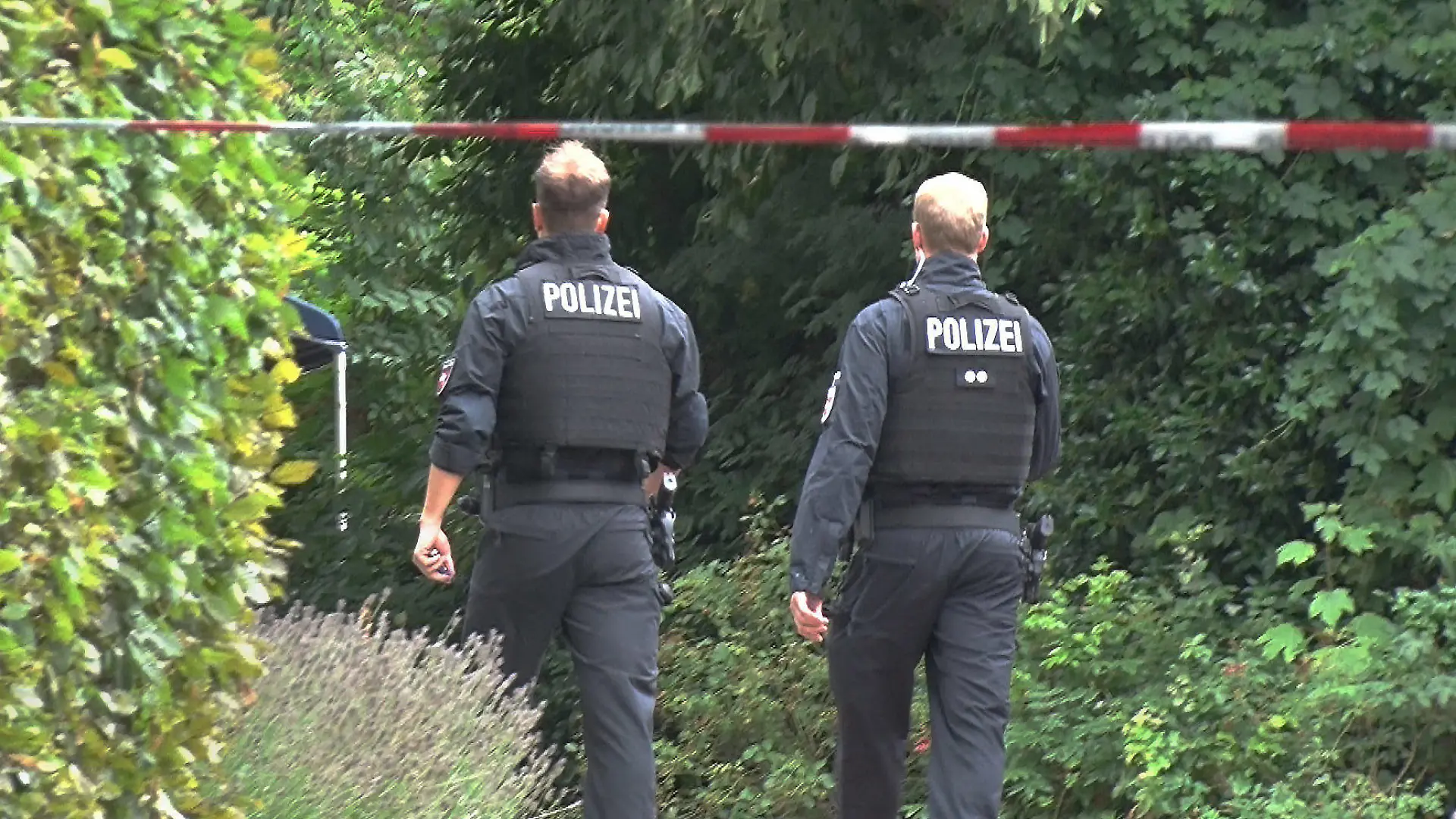 23.07.2023, Niedersachsen, Spelle: Zwei Polizisten gehen hinter einer Absperrung zu einem Gebüsch. Eine Spaziergängerin hat in einem Gebüsch in Spelle (Landkreis Emsland) einen toten Mann gefunden. Ein Fremdverschulden wird nicht ausgeschlossen, teilte die Polizei am Sonntagabend mit. Aus ermittlungstaktischen Gründen könne die Polizei aber derzeit nicht mehr zu dem Fund vom Mittag herausgeben, sagte eine Sprecherin in Lingen. Foto: -/NWM-TV/dpa +++ dpa-Bildfunk +++
